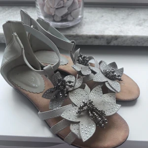 Beige sandaler med blommor strl 35 - Snygga beige sandaler med dekorativa blommor framtill, pärldetaljer och små silvriga nitar. Skorna har öppen tå, platt sula och dragkedja i hälen. Perfekta för varma dagar och ger en söt touch till din outfit.