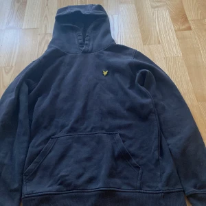 Svart hoodie från Lyle & Scott - Snygg svart hoodie från Lyle & Scott med klassisk gul logga på bröstet. Tröjan har huva, känguruficka och är tillverkad i mjuk bomull. Perfekt för chill dagar eller när du vill ha en enkel men stilren look.