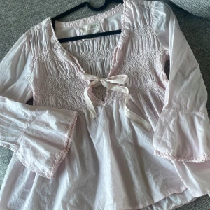 Ljusrosa blus från Odd Molly - Supersöt ljusrosa blus från Odd Molly med smock upptill, v-ringning och knytband framtill. Vida ärmar med spetsdetaljer och romantisk känsla. Perfekt för dig som gillar boho-vibe och mjuka pasteller. OPSS!! Står ingen storlek på men skulle säga att det hade passat en 10åring🫶🏻
