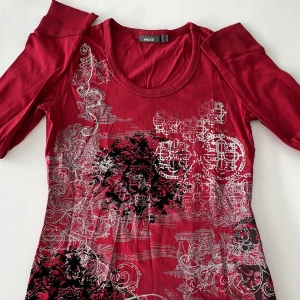 Röd topp med mönster från Mexx - Snygg röd topp från Mexx med coolt vitt och svart mönster framtill. Rund halsringning och trekvartsärm ger en modern vibe. Tunn och mjuk i bomull, perfekt för dig som gillar att sticka ut med detaljerade prints.