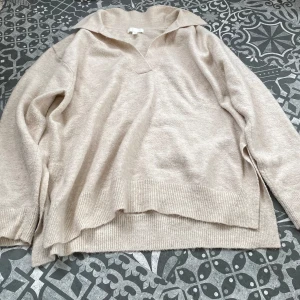 Beige stickad polotröja - Beige stickad polotröja med v-ringning och bred krage. Tröjan har långa ärmar, ribbade muddar och en avslappnad passform med slits i sidorna. ☀️
