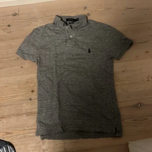 Grå pikétröja från Polo Ralph Lauren - Snygg grå pikétröja från Polo Ralph Lauren med klassisk krage, knappar vid halsen och den ikoniska broderade loggan på bröstet. T-shirten är kortärmad och har en slim fit pass form, endast använd några få gånger pågrund av att den inte passade.