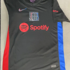 FC Barcelona svart Nike fotbollströja S - Svart FC Barcelona fotbollströja från Nike med röda och blå sidopaneler. Tryck med Spotify-logga på bröstet och UNHCR ACNUR på ryggen. Klassisk klubbsköld och Nike Swoosh framtill. Tillverkad i lätt och ventilerande material.
