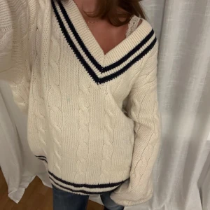 Vit kabelstickad v-ringad ullröja - En oversized vit kabelstickad ulltröja med v-ringning och mörkblå ränder vid halsen och nederkanten. Knappt använd, men tröjan har dock en liten blå fläck som troligtvis kommit från de blåa ränderna. (syns på sista bilden)