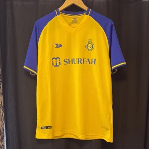 Al Nassr FC gul/blå matchtröja L - Gul och blå officiell Al Nassr FC fotbollströja från säsongen 2022-2023, tillverkad av Duneus Sportswear. Tröjan har korta ärmar, V-ringning och klubbmärke på bröstet. Materialet är lätt och andas, perfekt för match eller träning. Köpt i saudi Arabien.