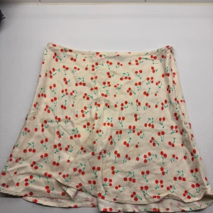 Mini skirt med körsbär från Zara - Supersöt vit kortkjol från Zara med färgglatt körsbärsmönster. Kjolen har en lätt utsvängd form och dragkedja i sidan. Perfekt för dig som vill ha en lekfull och somrig vibe i garderoben.