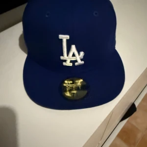 Blå LA Dodgers keps från New Era - Snygg blå LA Dodgers keps från New Era med klassisk vit LA-logga framtill och MLB-logga bak. Modellen är 59FIFTY med rak skärm och broderade ventilationshål. Perfekt för dig som vill ha en sportig och stilren look.