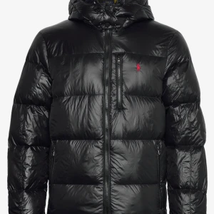 Svart pufferjacka från Polo Ralph Lauren - Snygg svart pufferjacka från Polo Ralph Lauren med röd logga på bröstet. Jackan har huva, dragkedjor på fickor och bröst, samt quiltad design. Perfekt för kalla dagar och riktigt skön att bära. Klassisk look som funkar till allt.
