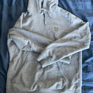 Grå hoodie från Lyle & Scott - Säljer en grå hoodie från Lyle & Scott med klassisk gul logga på bröstet. Tröjan har huva med snörning och en stor magficka. Perfekt för chill dagar och enkel att matcha med jeans eller joggers.