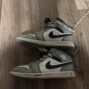Nike Air Jordan 1 grå och vita sneakers - Nike Air Jordan 1 sneakers i grått och vitt med svarta detaljer och klassisk swoosh-logga. Skorna har snörning och hög siluett, samt ikonisk Air Jordan-logga på sidan. Perfekta för dig som gillar streetstyle och vill sticka ut med en tidlös modell.