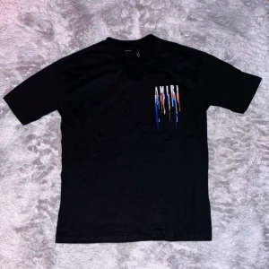 Svart AMIRI t-shirt med färgstänk - Svart t-shirt från AMIRI med stort vitt logotryck och färgstänk i blått, rött och vitt på både bröstet och ryggen. Sprillans ny, säljer pga för stor för mig. Hör av dig vid intresse 👋