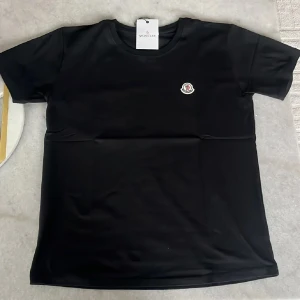 Svart Moncler t-shirt med logga - Svart t-shirt från Moncler med klassisk rund hals och liten broderad logga på bröstet. Enkel och stilren design som passar perfekt till jeans eller shorts. Tillverkad i mjuk bomull för en skön känsla hela dagen.
