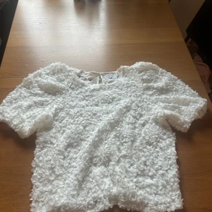 Vit blommig blus med 3D-struktur - Supersöt vit blus med kort ärm och 3D-blommor över hela tyget. Blusen har rund halsringning och en liten knapp i nacken. Perfekt för dig som vill sticka ut med en romantisk och trendig look.