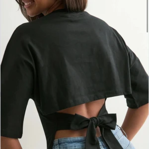 Nelly open back tie teee - Säljer en svart topp med halvlånga ärmar och snygg omlottdesign. Toppen har öppen rygg med breda band som knyts i en stor rosett bak. Figurnära passform och rund halsringning. Perfekt för dig som vill ha en trendig och unik look❣️
