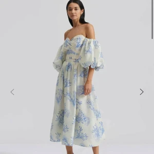 By Malina offshoulder klänning blå/vit - Superfin offshoulder klänning från By Malina i ljus bas med blått korallmönster. Klänningen har puffiga ärmar, omlottkänsla upptill och en längre kjol. Materialmix av tencel, bomull och linne, fodrad med bomull. Perfekt för sommarens alla tillfällen. Endast testad.