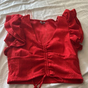 Röd blus med volangärm från FP Sister - Superfin röd blus från FP Sister med v-ringning, volangärmar och snygg snörning framtill. Blusen har broderade detaljer och är croppad, perfekt för dig som vill sticka ut lite extra. Luftigt material som passar till varma dagar.