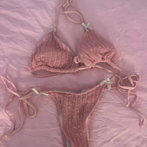 Rosa ribbad bikini med rosetter - Supersöt rosa bikini med ribbad struktur och små vita rosetter på både överdel och underdel. Bikinin har knyt i sidorna och runt nacken för perfekt passform. Materialet är mjukt och stretchigt, perfekt för sommardagar på stranden.
