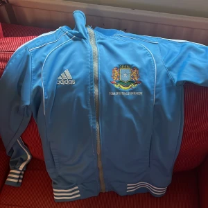Adidas Somalia träningsjacka blå - Blå träningsjacka från Adidas med Somalia Football Federation-logga på bröstet och 'SOMALIA' broderat på ryggen. Jackan har vita ränder på ärmarna, dragkedja framtill och ribbade muddar. Perfekt för dig som gillar fotboll och vill sticka ut med en unik jacka.