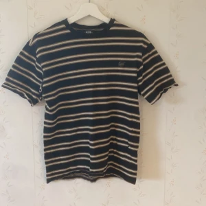 Randig t-shirt från Sweet SKTBS - Snygg randig t-shirt från Sweet SKTBS i svart, beige och brunt. Klassisk passform med korta ärmar och broderad logga på bröstet. Perfekt för dig som gillar streetwear och vill ha en stilren look. Gjord i mjuk bomull för skön känsla hela dagen.