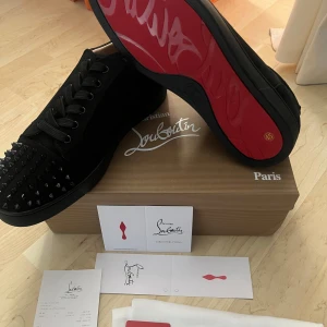 Christian Louboutin svarta sneakers med nitar - Svarta sneakers från Christian Louboutin i mocka med ikonisk röd sula och svarta nitar på tån. Skorna har snörning och en platt sula, vilket ger en edgy och lyxig vibe. Perfekta för dig som vill sticka ut med exklusiva detaljer och streetstyle-känsla.