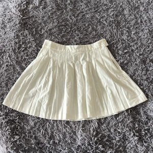 Vit plisserad kjol från FB Sister - Superfin vit plisserad kortkjol från FB Sister i storlek L. Kjolen har bred linning, dragkedja och knapp i sidan för enkel påklädning. Perfekt för en fräsch och stilren look, passar till sneakers eller boots.