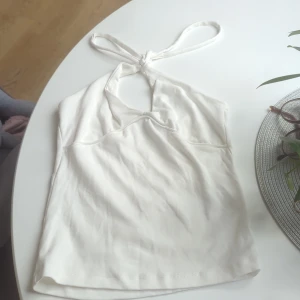 Vit halterneck linnetopp - Snygg vit linnetopp med halterneck och knyt i nacken. Toppen har en lätt v-ringad skärning och är ärmlös, perfekt för varma dagar. Enkel och stilren design som passar till många olika outfits.