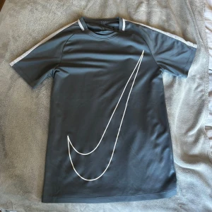 Grå Nike t-shirt med vit logga - Snygg grå t-shirt från Nike med stor vit swoosh-logga över framsidan. T-shirten har korta ärmar och vita detaljer längs axlarna. Perfekt för dig som gillar sportig och clean stil.