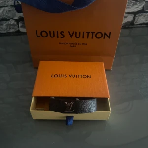 Louis Vuitton svart armband monogram - Snyggt svart armband från Louis Vuitton i klassiskt monogrammönster med LV-logga i silver. Tillverkat i präglat skinn och levereras i originalask med tillhörande kort. Perfekt accessoar för dig som gillar exklusiva detaljer och ikonisk design. Pris kan diskuteras