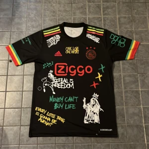 Ajax x Bob Marley svart fotbollströja - Unik svart Ajax fotbollströja från Adidas med Bob Marley-tema. Flera färgglada tryck och texter, röd Ziggo-logga, Ajax-emblem och graffiti-inspirerade detaljer. Ärmarna har röd, gul och grön rand. Materialet är lätt och andas, perfekt för fotboll eller streetwear.