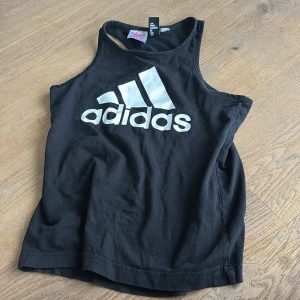 Svart Adidas linnetopp med logga - Svart linnetopp från Adidas med stor vit logga framtill. Modellen är ärmlös med brottarrygg och tillverkad i mjuk bomull, perfekt för träning eller chill dagar. Enkel och sportig design som är lätt att matcha med andra plagg.
