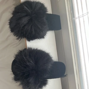Svarta fluffiga sandaler med päls - Supermjuka svarta sandaler med stor fluffig pälsdetalj över foten. Perfekta för att chilla hemma eller ge din outfit en extra touch. Platt sula och öppen tå för maximal komfort.