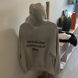 Grå hoodie från Acne Studios - Säljer en grå hoodie från Acne Studios med tryck på ryggen där det står 'Acne Studios STOCKHOLM 1996'. Klassisk passform med huva och långa ärmar samt sliten design. Perfekt för dig som gillar stilren och enkel streetwear. Den är i strl XS, men är stor i storleken. Nypriset på den är 5300 kr. 