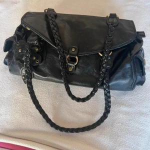 Svart axelväska med flätade handtag - Snygg svart axelväska med två flätade handtag och detaljer i mässingsfärgad metall. Väskan har lock med tryckknapp, dragkedja upptill och flera nitar som ger en cool look. Perfekt storlek för allt du behöver ha med dig. Den är stor!
