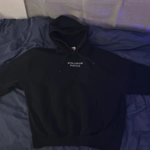 Svart hoodie med tryck från Fortysix - Säljer en svart hoodie med texten 'WORLDWIDE SERVICE' framtill och stort gult 'FORTYSIX'-tryck på ryggen. Klassisk huva. Perfekt för en avslappnad streetwear-look.