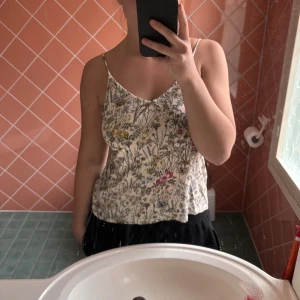 Blommig linnetopp med tunna axelband - Säljer en söt topp med tunna axelband och blommigt mönster i flera färger. Toppen är vit i grunden med detaljer i gult, rosa, lila och grönt. 