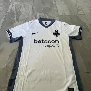 Inter Milan vit fotbollströja Nike - Säljer en vit Inter Milan fotbollströja från Nike med grå detaljer och svarta loggor. Tröjan har korta ärmar, V-ringad krage och klubbmärke samt sponsortryck på bröstet. Perfekt för dig som älskar fotboll och vill visa ditt stöd för Inter.