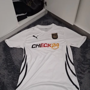 Vit Tyskland fotbollströja från Puma - Vit fotbollströja från Puma med Tysklands örn och texten 'DEUTSCHLAND' på bröstet. Svarta detaljer längs sidorna, svart Puma-logga och sponsortryck 'CHECK24' i svart, rött och gult. Tillverkad i polyester.