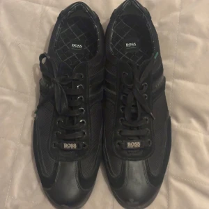 Svarta sneakers från Hugo Boss - Svarta sneakers från Hugo Boss med klassisk design. Skorna har snörning, rund tå och är tillverkade i en mix av skinn och syntetmaterial. Skriv för fler frågor eller bilder i chatten tack!