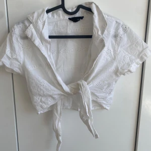 Vit croppad blus med knyt - Säljer en vit croppad blus med korta ärmar och knyt framtill. Blusen har broderade detaljer i form av små hålmönster och en klassisk krage. Perfekt för varma dagar och ger en fräsch sommarkänsla. Kan verkligen ge något extra till outfiten i sommar!
