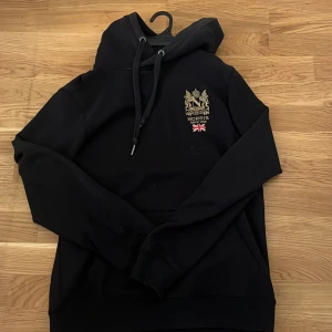 Svart hoodie från Morris - Säljer en svart hoodie från Morris med broderad logga och brittisk flagga på bröstet. Tröjan har huva med dragsko och en klassisk känguruficka framtill. Perfekt för en stilren och avslappnad look.