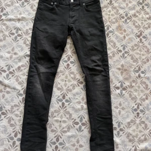 Svarta Nudie Jeans - Säljer ett par svarta jeans från Nudie Jeans med klassisk femficksdesign och broderad bakficka. Jeansen har normal passform och raka ben. Perfekta för dig som gillar stilrena och tidlösa jeans.