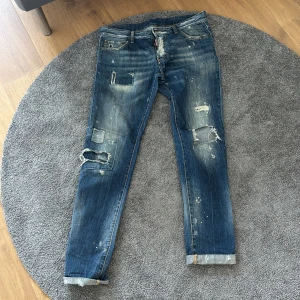 Blå slitna jeans från DSQUARED2 - Säljer ett par blå jeans från DSQUARED2 med slitningar, hål och lagningar för en riktigt rå look. Jeansen har klassisk femficksdesign, uppvikta benslut och tydliga slitdetaljer framtill och baktill. Perfekta för dig som gillar streetstyle. Skriv ifall ni vill ha mer information😀!