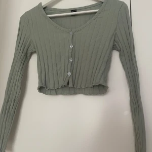 Ljusgrå croppad kofta med knappar - Säljer en ljusgrön croppad kofta. Tröjan är ribbad med långa ärmar och har knappar framtill. Perfekt att styla över ett linne eller t-shirt för en trendig look.