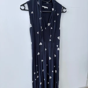 Mörkblå klänning med hjärtmönster från mint & berry - Supersöt mörkblå klänning från mint & berry med vita hjärtan över hela tyget. Ärmlös modell med v-ringning och knytband vid halsen. Perfekt för dig som gillar unika detaljer och lekfulla mönster.