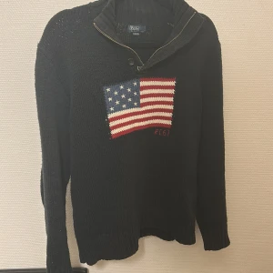 stickad half zip Ralph Lauren - Svart stickad tröja från Polo Ralph Lauren med en stor amerikansk flagga broderad på bröstet. Tröjan har hög krage med dragkedja och knapp, samt långa ärmar. Riktigt snygg och sällsynt i perfekt skick. Perfekt för dig som gillar klassisk och sportig stil. Passar dig mellan 175-185. Skriv vid frågor