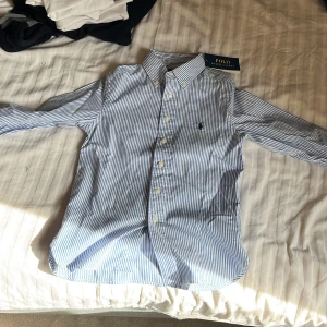 Blåvit randig skjorta från Polo Ralph Lauren - Säljer en klassisk blåvit randig skjorta från Polo Ralph Lauren med button-down krage och broderad logga på bröstet. Skjortan har långa ärmar och knäppning framtill. Perfekt för en stilren look.
