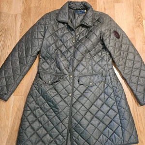 Quiltad grön kappa från Polo Ralph Lauren - Säljer en lång, quiltad grön kappa från Polo Ralph Lauren. Jackan har klassisk krage, tryckknappar framtill och två stora fickor med lock. Perfekt för kyligare dagar och har en stilren look med diskret logotyp på ärmen. Kappan är vattenavvisande. Inköpt på Pondus för 4490 kr.
