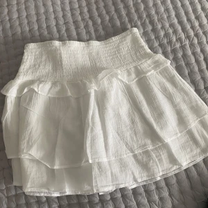 Vit volangkjol från Gina Tricot - Supersöt vit kjol från Gina Tricot med volanger och smock i midjan. Kjolen är kort och har en luftig känsla, perfekt för varma dagar. Passar dig som gillar en feminin och trendig stil.