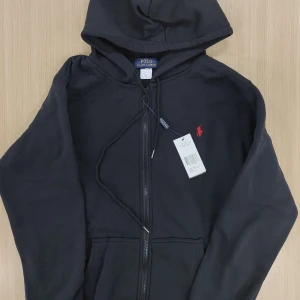 Svart hoodie från Polo Ralph Lauren - Svart hoodie från Polo Ralph Lauren med dragkedja framtill och röd broderad logga på bröstet. Tröjan har huva med snörning och två fickor framtill. Perfekt för en avslappnad stil.  Går ner i pris vid snabb affär