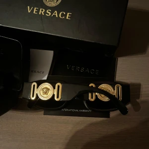 Svarta solglasögon från Versace med guldiga detaljer - Exklusiva svarta solglasögon från Versace med ikoniska guldiga Medusa-detaljer på skalmarna. Glasögonen har en klassisk, rektangulär form och levereras med originaletui och tillbehör såsom verifieringskod, putsduk osv. Hör av er vid funderingar. Svarar ofta snabbt🫶